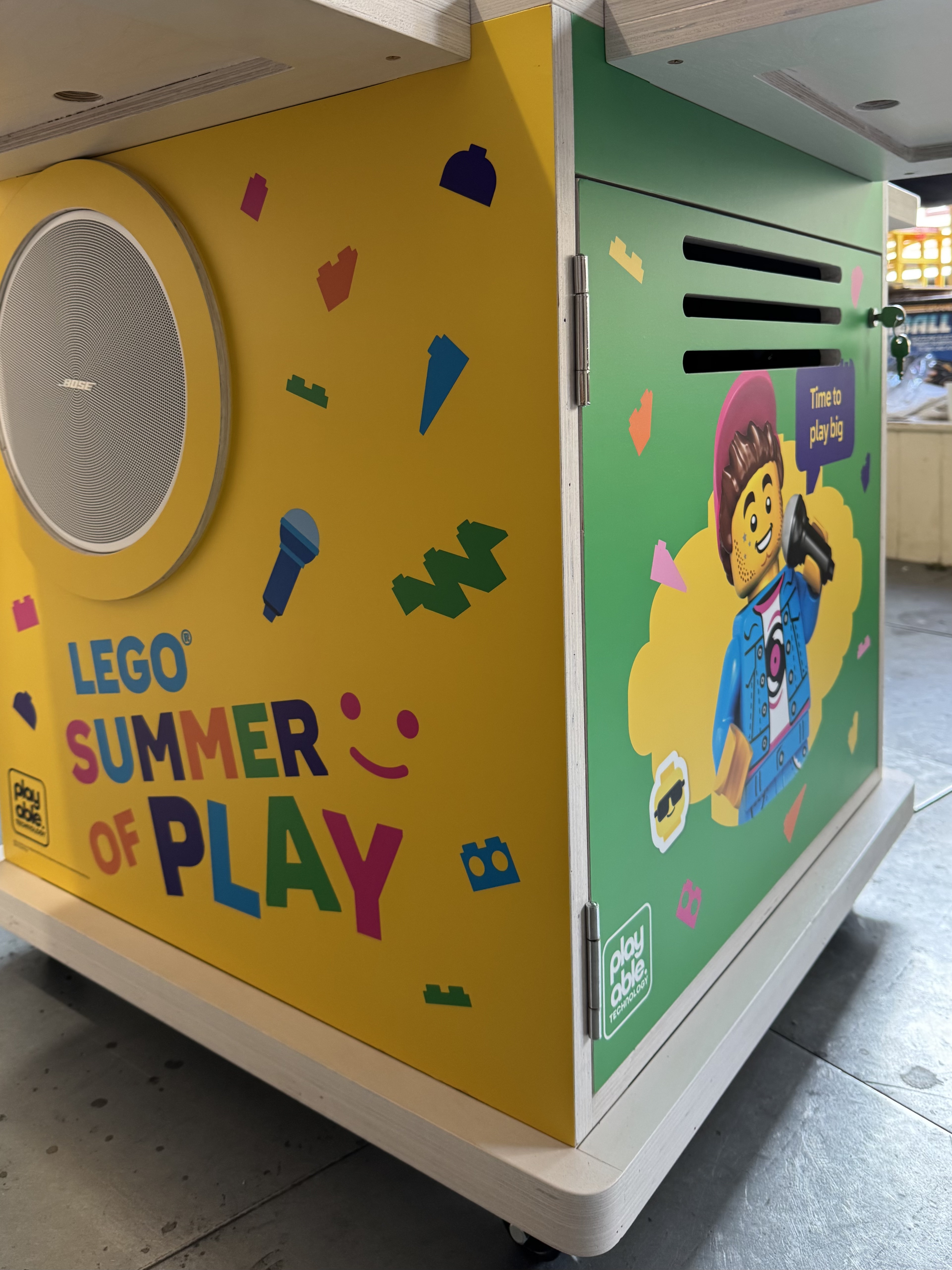 A finished BeatBricks unit on castors in the workshop, showing two sides of its LEGO Summer of Play livery, the yellow speaker panel and the green minifigure panel.