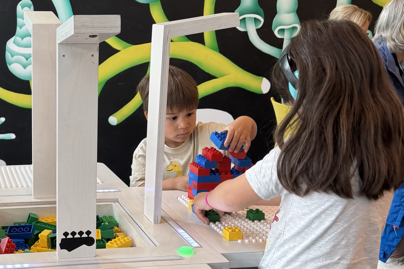 A boy builds a tall red and blue brick tower on the BeatBlocks Live baseplate while another child reaches in, at V&A Dundee.
