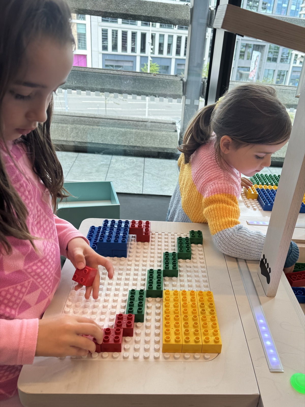 Two girls build with coloured bricks at BeatBlocks Live by the studio window at V&A Dundee.
