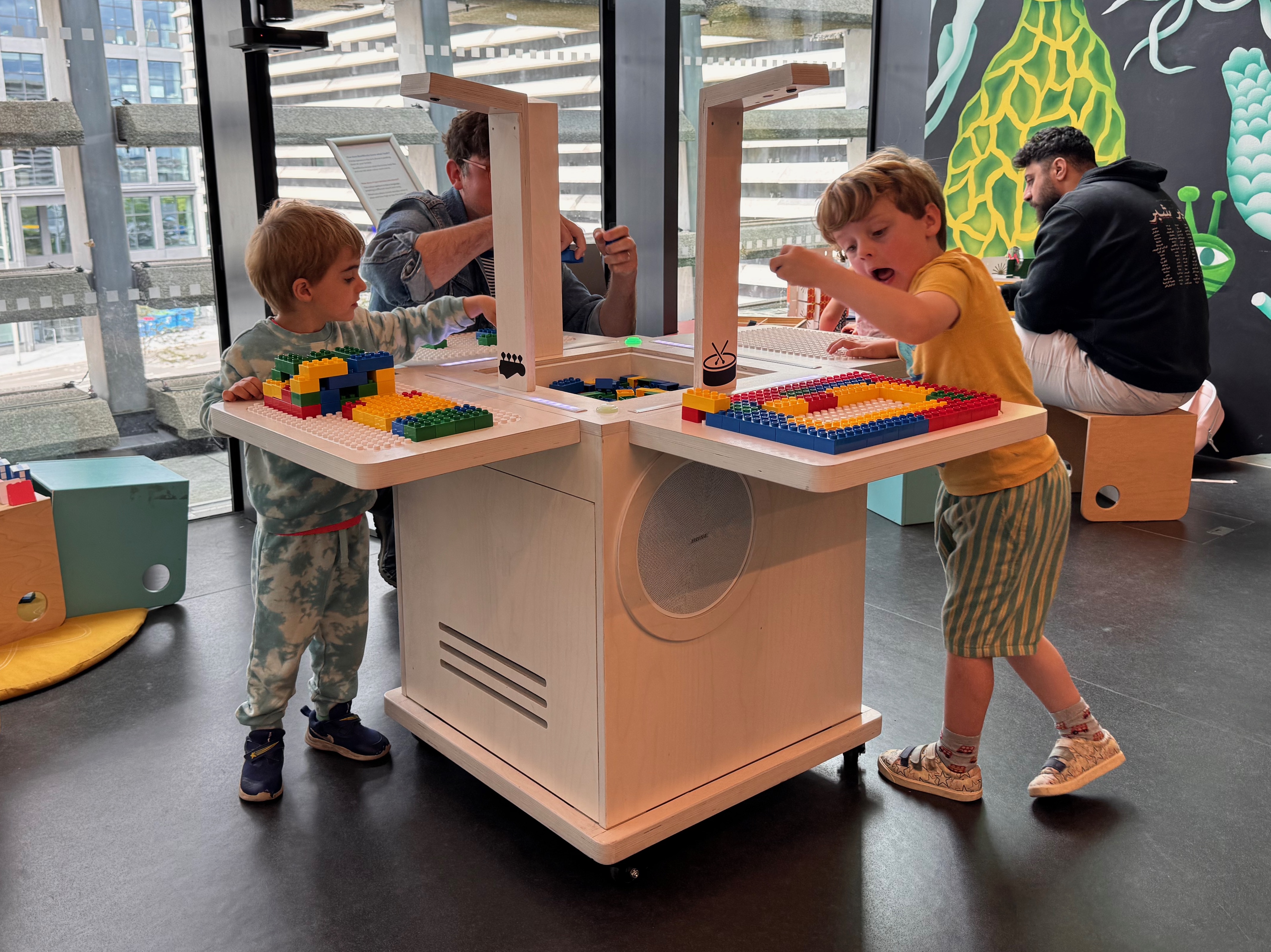 Two young boys build patterns of coloured bricks on adjacent baseplates of the BeatBlocks Live unit at V&A Dundee, an adult playing at the far side and a green mural behind.