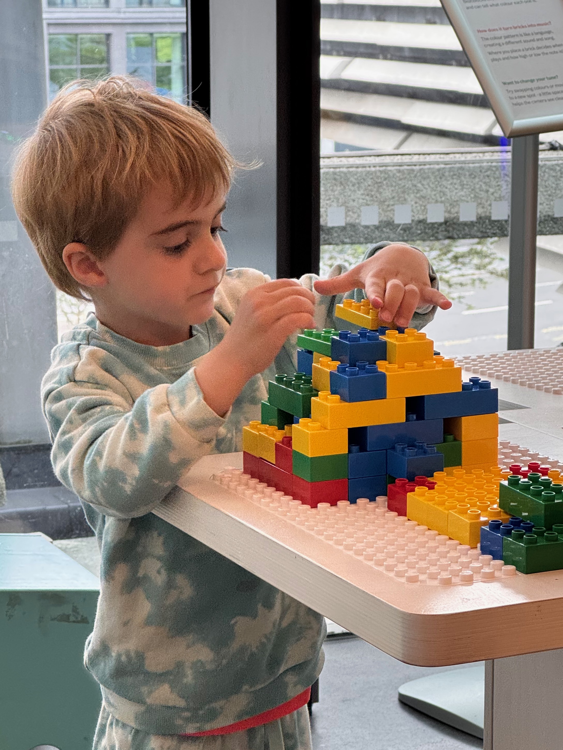 A young boy carefully stacks a tall tower of coloured bricks on a BeatBlocks Live baseplate at V&A Dundee, an instruction panel behind him.