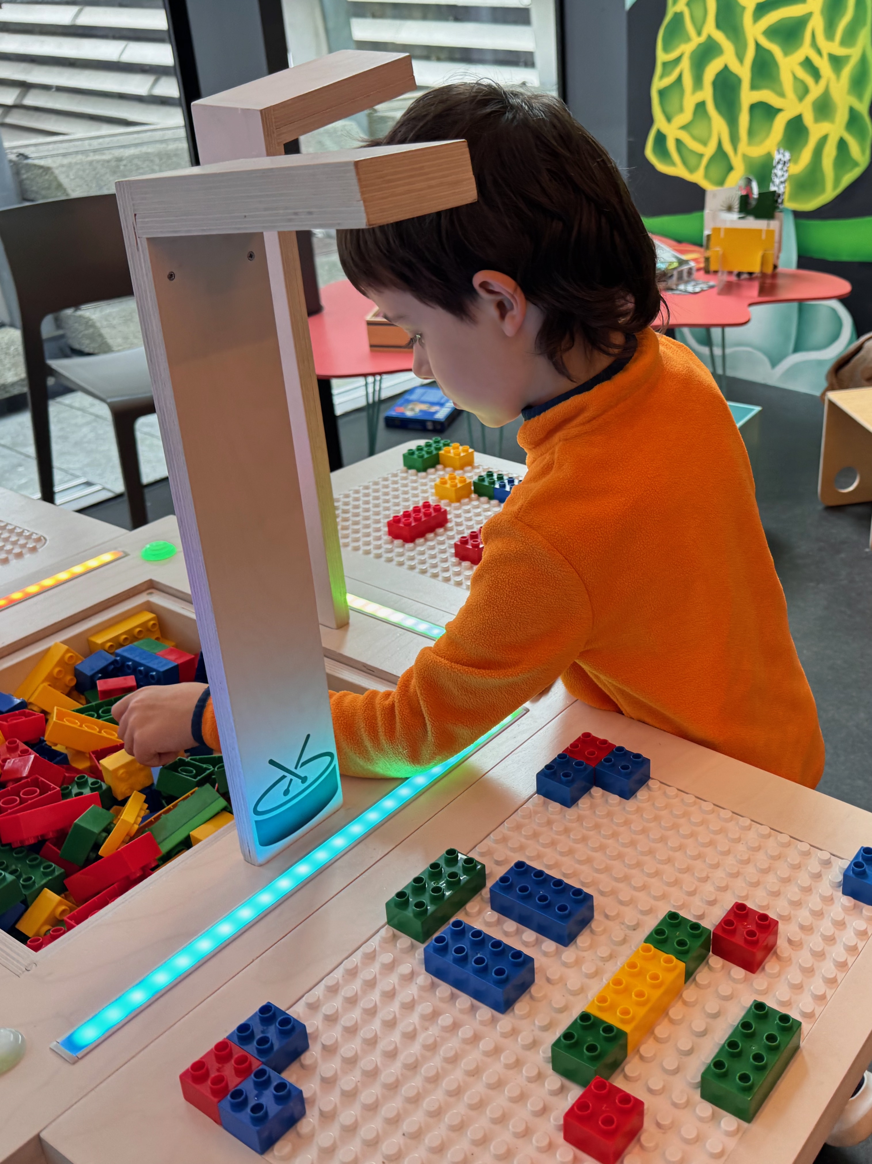 A boy in an orange top reaches into the brick bin at the BeatBlocks Live unit at V&A Dundee, the lit camera column in front and a part-built baseplate in the foreground.