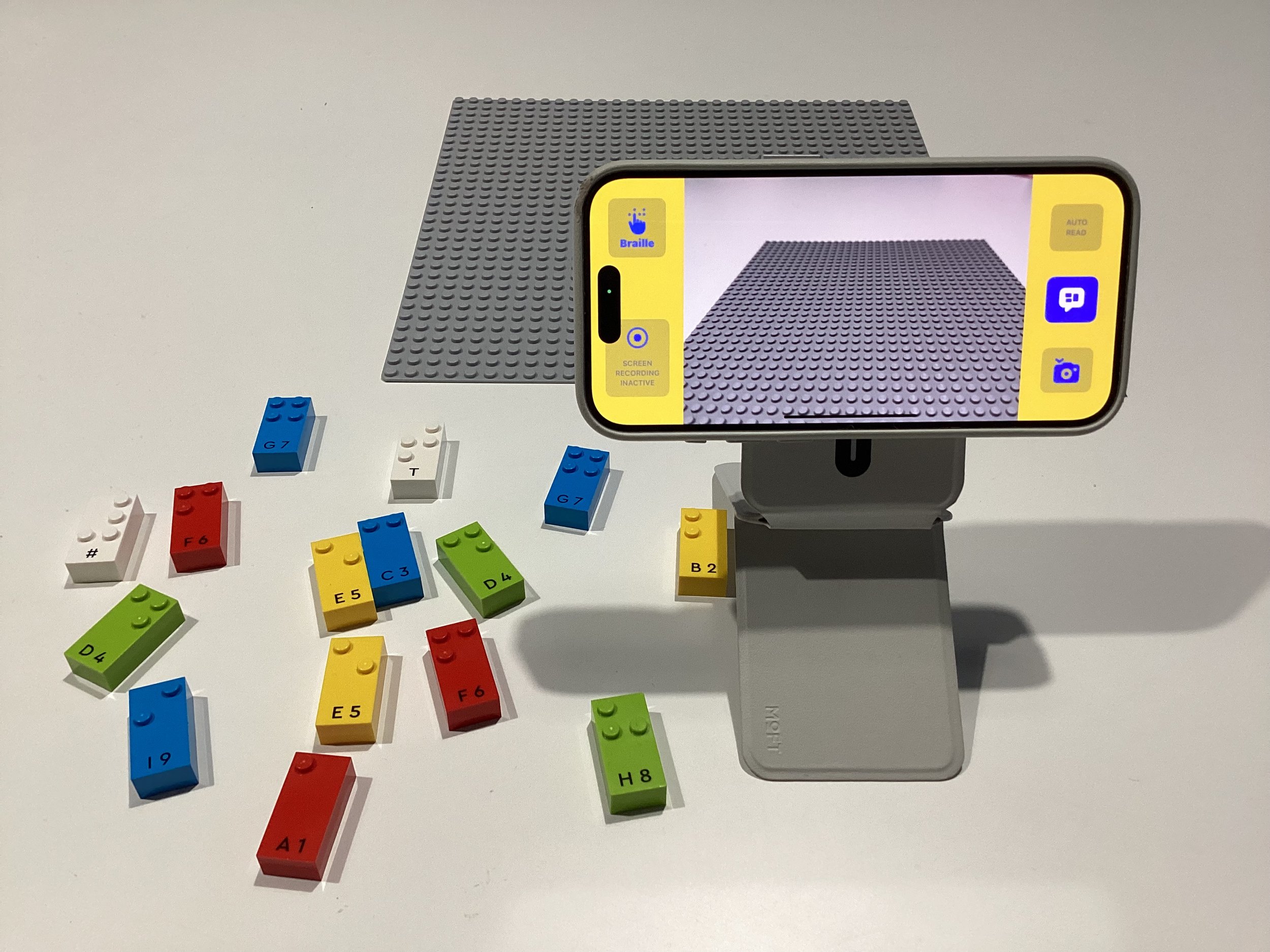 An iPhone on a stand runs BrailleBlocks in Braille mode, facing a grey LEGO baseplate, with lettered LEGO Braille bricks laid out on the table.