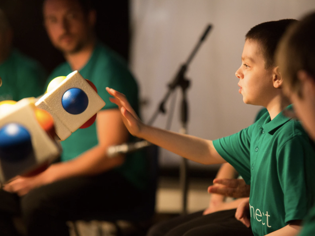 A young boy in a green shirt reaches out to play a Skoog, a soft cube instrument with coloured buttons, during a performance.