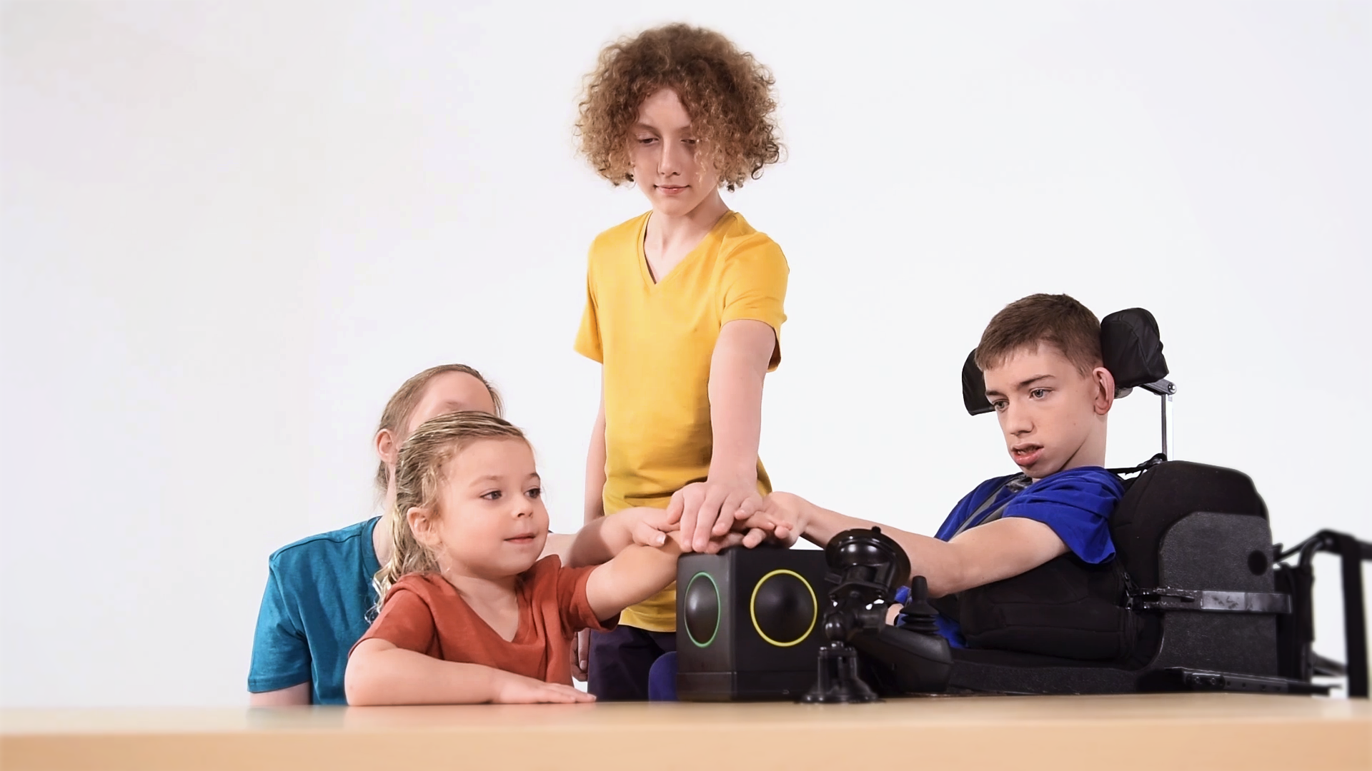 Four children, one using a powered wheelchair, playing a single Skoog together at a table — reaching in from seated and standing positions.